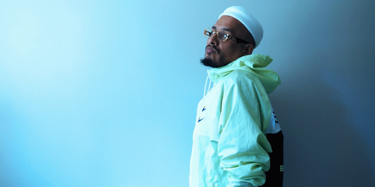 Bocacloja facing the left with head leaning towards the lens. Wearing glasses, a white beanie, and track jacket. Background is blue.