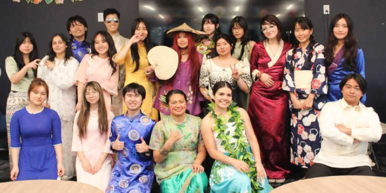19 people standing in a group smiling at the camera wearing various Asian and Pacific Islander traditional clothing
