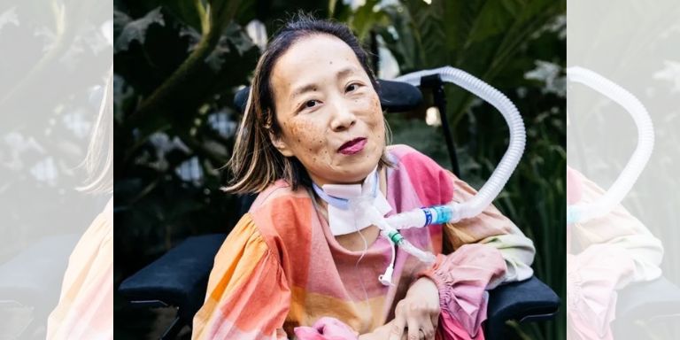 An Asian American Disabled woman sits in a power chair with a tracheostomy with a tube attached to her ventilator. She is wearing pink and orange tie dye, and is smiling at the camera. 