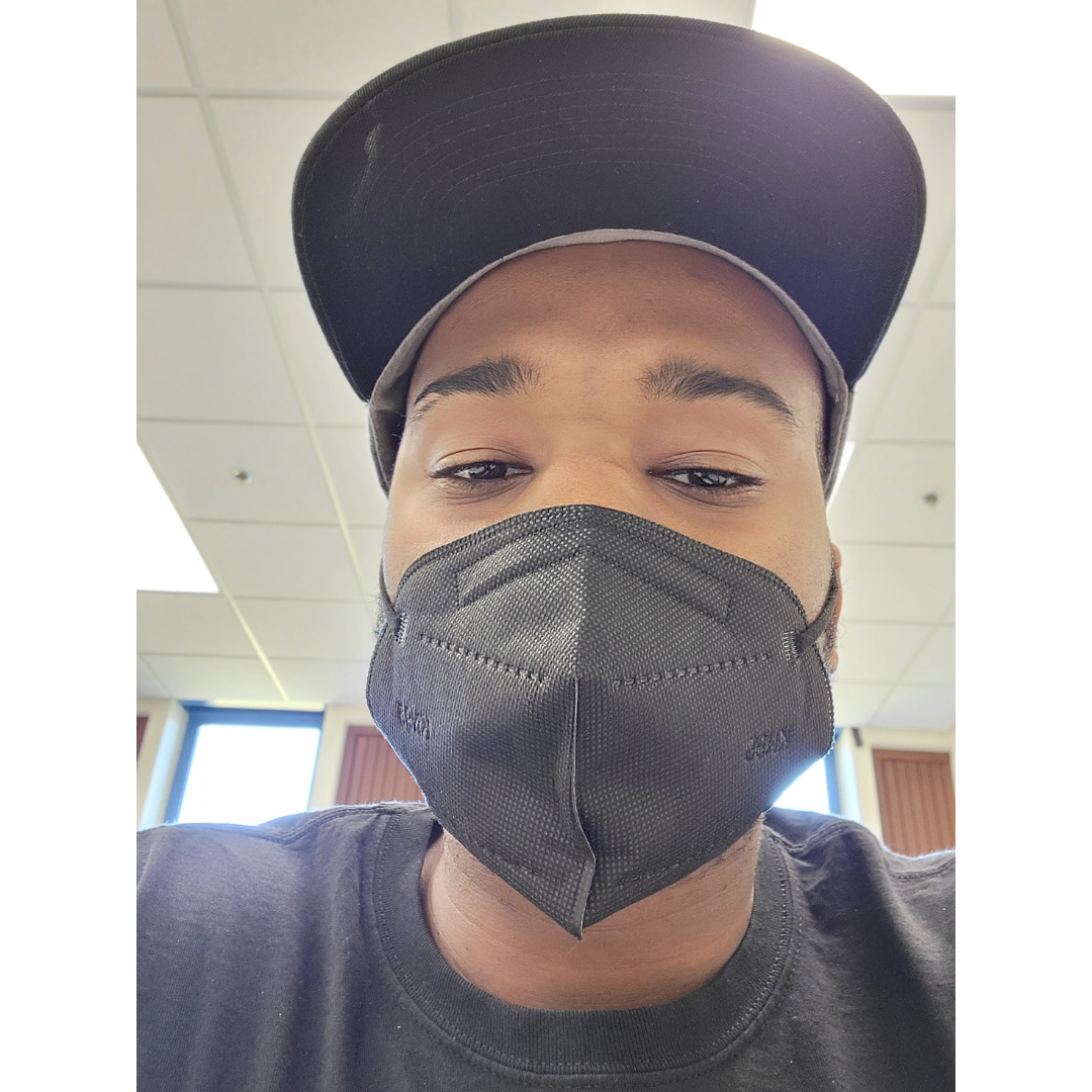 image of a person looking at the camera. They are wearing a gray mask covering most of their lower face and they are also wearing a hat and gray shirt.