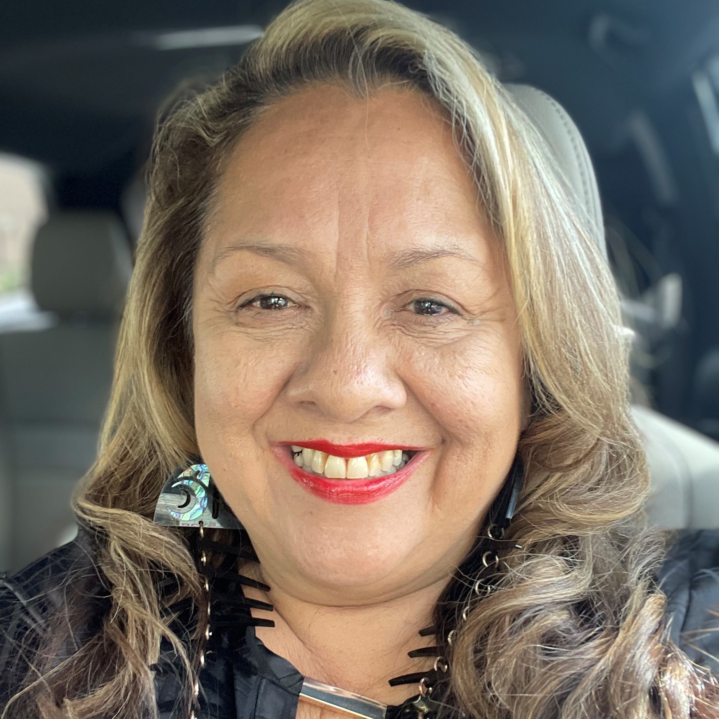 Head shot of Lori Laiwa Thomas. She is smiling at the camera. Her hair is long, brown, and curled with blond highlights. She is wearing red lipstick and abalone fish earrings and a black top.