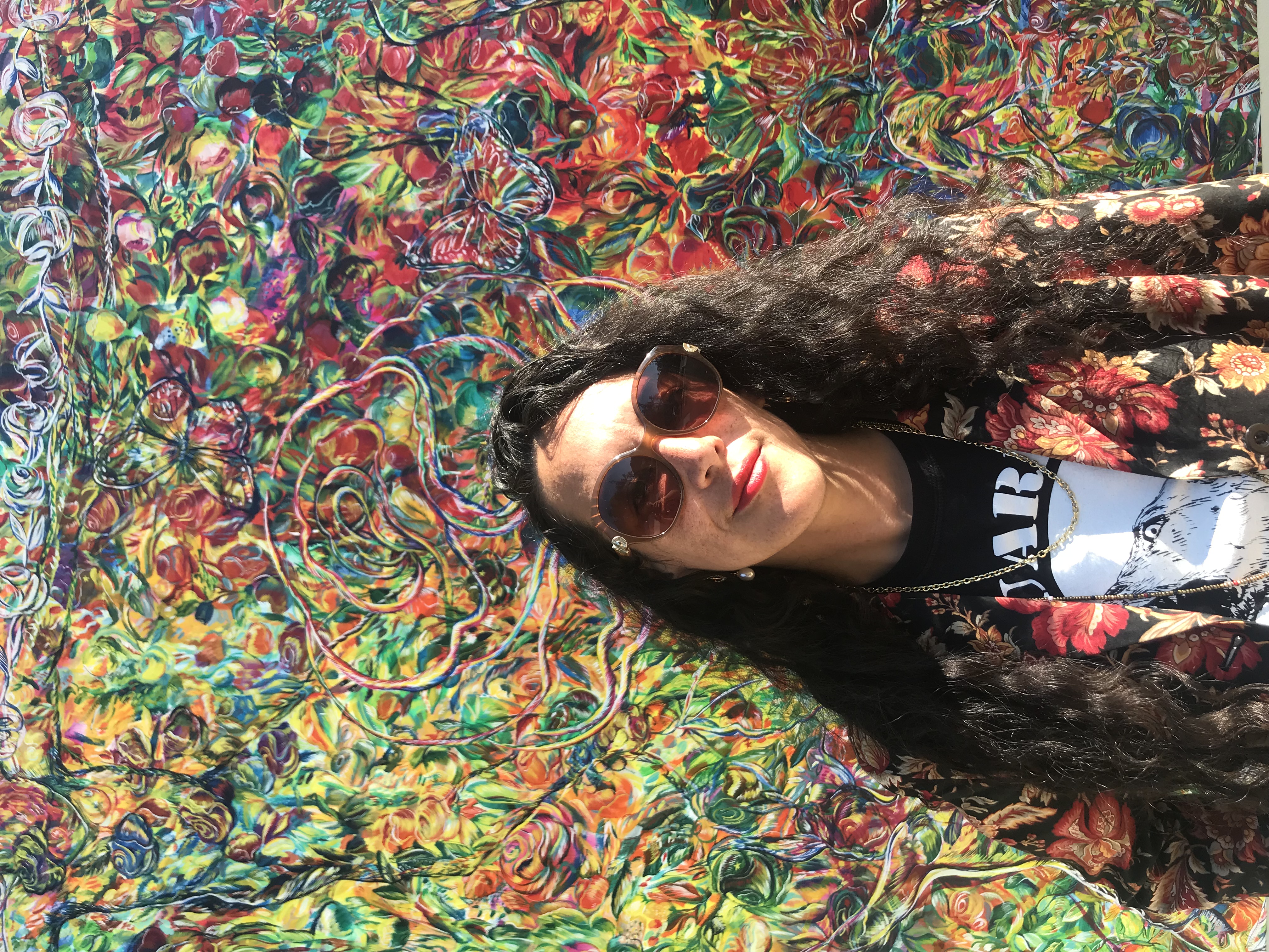 picture of Maria de los Angeles, long curly hair, sunglasses on, looking at the camera smiling. Standing infront of a mural full of colors and free formed lines