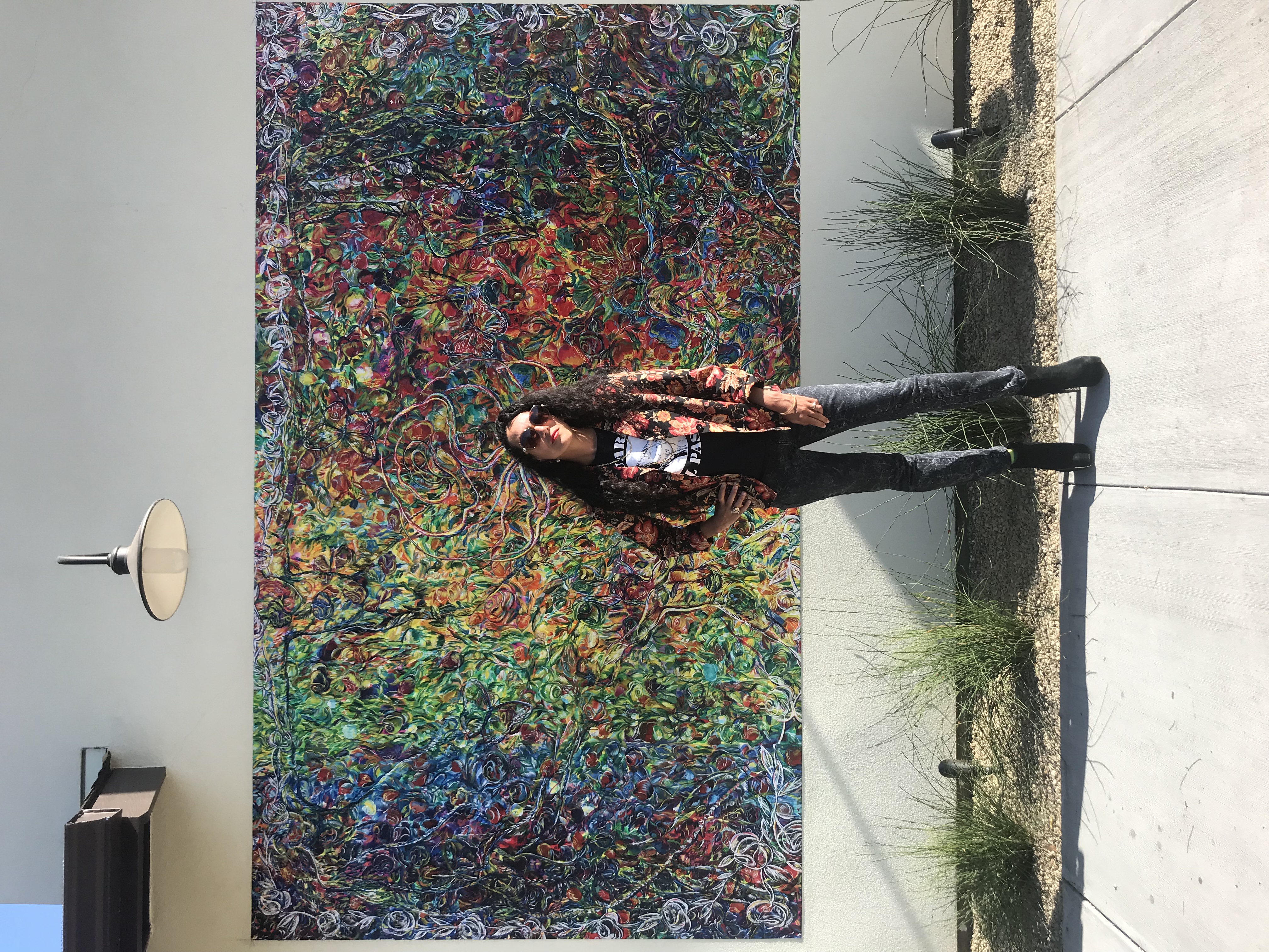 picture of Maria de los Angeles from far away, long curly hair, sunglasses on, looking at the camera smiling. Standing infront of a mural full of colors and free formed lines