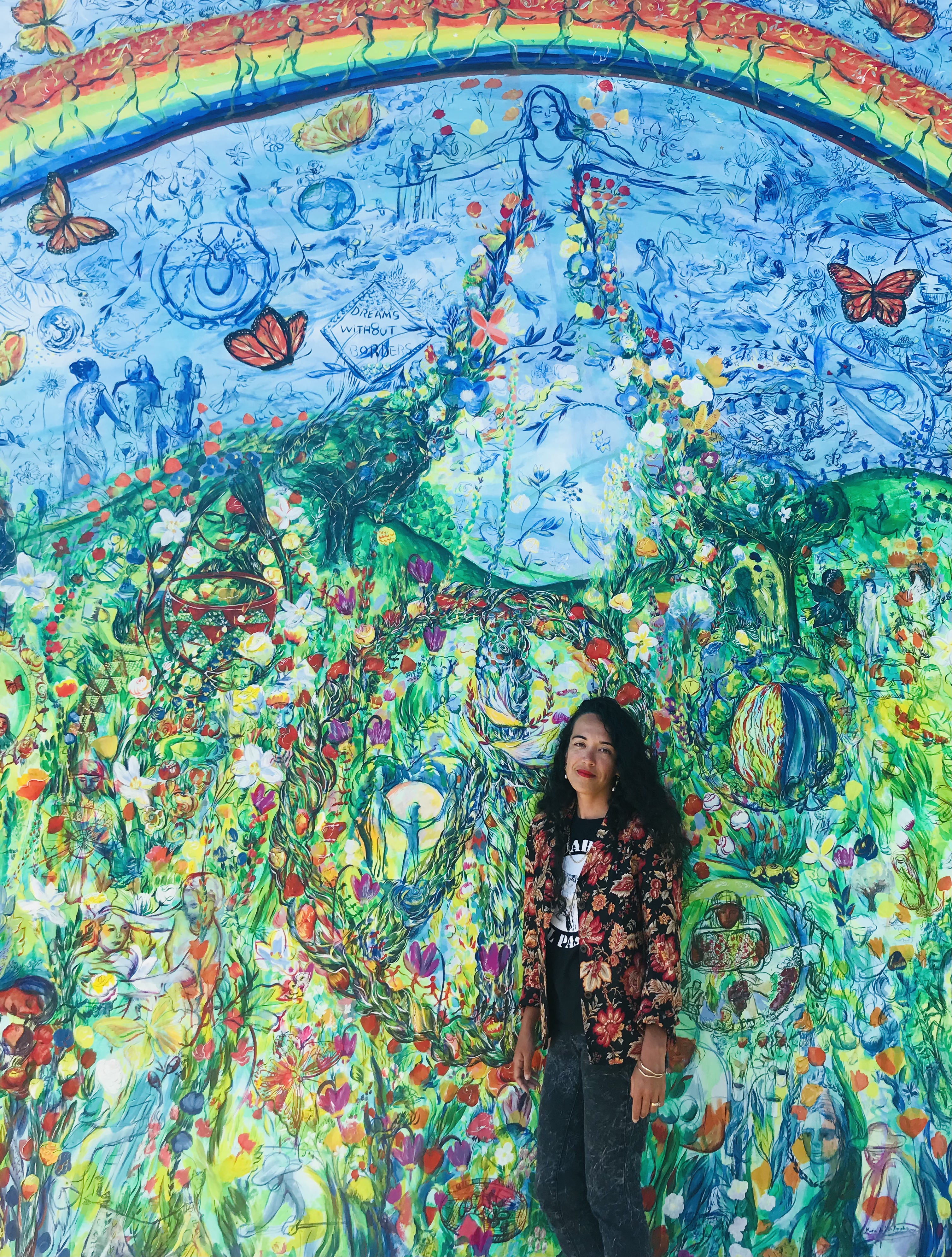 Picture of Maria de los Angeles, long curly hair, looking at the camera, standing in front of a mural of a person in a dress under a rainbow and standing over a hill. Other visuals are embedded within the art. 