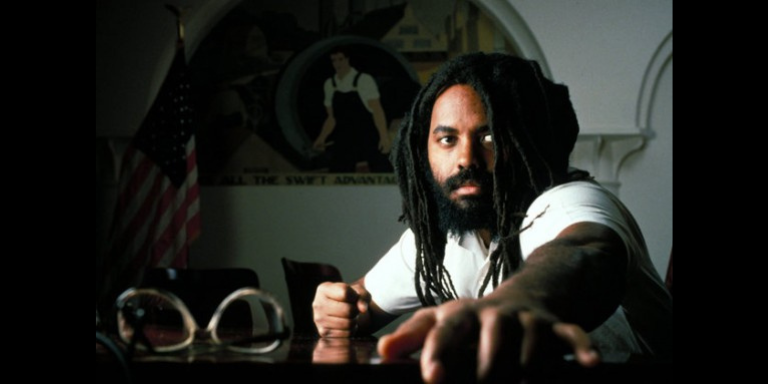 picture of Mumia sitting at a table with his glasses on the table and in the dark background the US flag