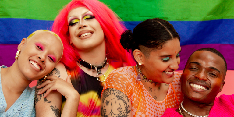 Four people smiling looking at the camera and at each other. They are wearing bright makeup and clothing with a pride flag in the background