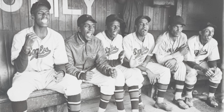 Black and White photograph of 6 Black baseball players sitting on the bench highly animated watching the game