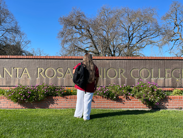 A student's back facing the camera. They have a black backpack hanging on one shoulder. They have white pants and red top. Their long hair falls down thier back. They are facing a brick and metal sign that reads "Santa Rosa Junior College" with green grass and purple flowers around it. 