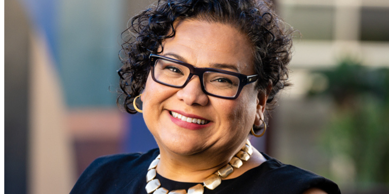 picture of Dr. Garcia, has short curly hair, is smiling at the camera. Wearing a bulky gold necklace over a black shirt.