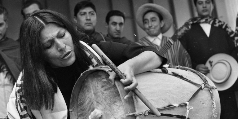 Black and Gray picture of mercedes sosa singing and playing a drum. In the background, which is out of focus, there are 5 people behind her watching her or looking elsewhere.
