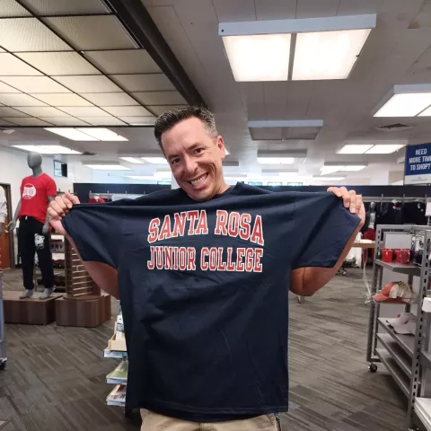 Big Cheesin smile on a happy go lucky dude who is holding up a Santa Rosa Junior College T-shirt inside of the Santa Rosa Junior College bookstore
