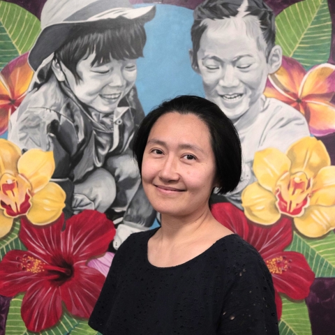 Picture of a person smiling at the camera with short black hair and a black blouse. They are standing in front of a mural of two children with flowers around them