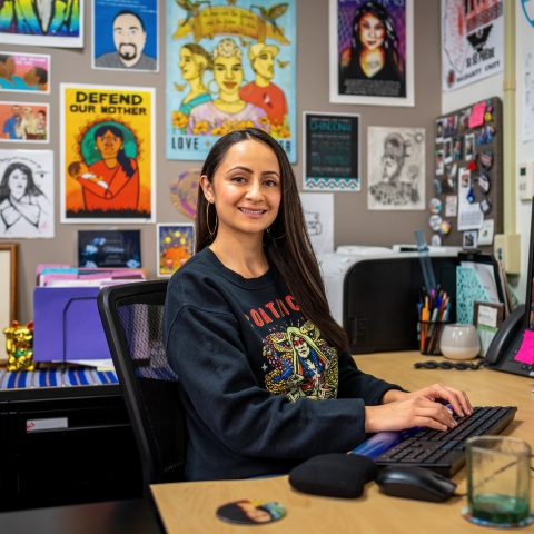 image of a Chicana with long hair sitting in front of a desk with colorful posters behind them